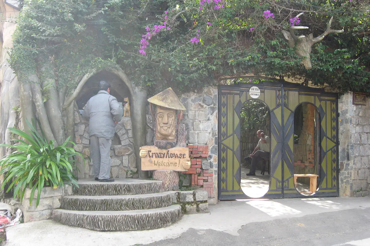 The entrance of Hang Nga Guesthouse (Crazy House) in Da Lat, with stone steps, a carved wooden figure, a welcome sign and bougainvillea above