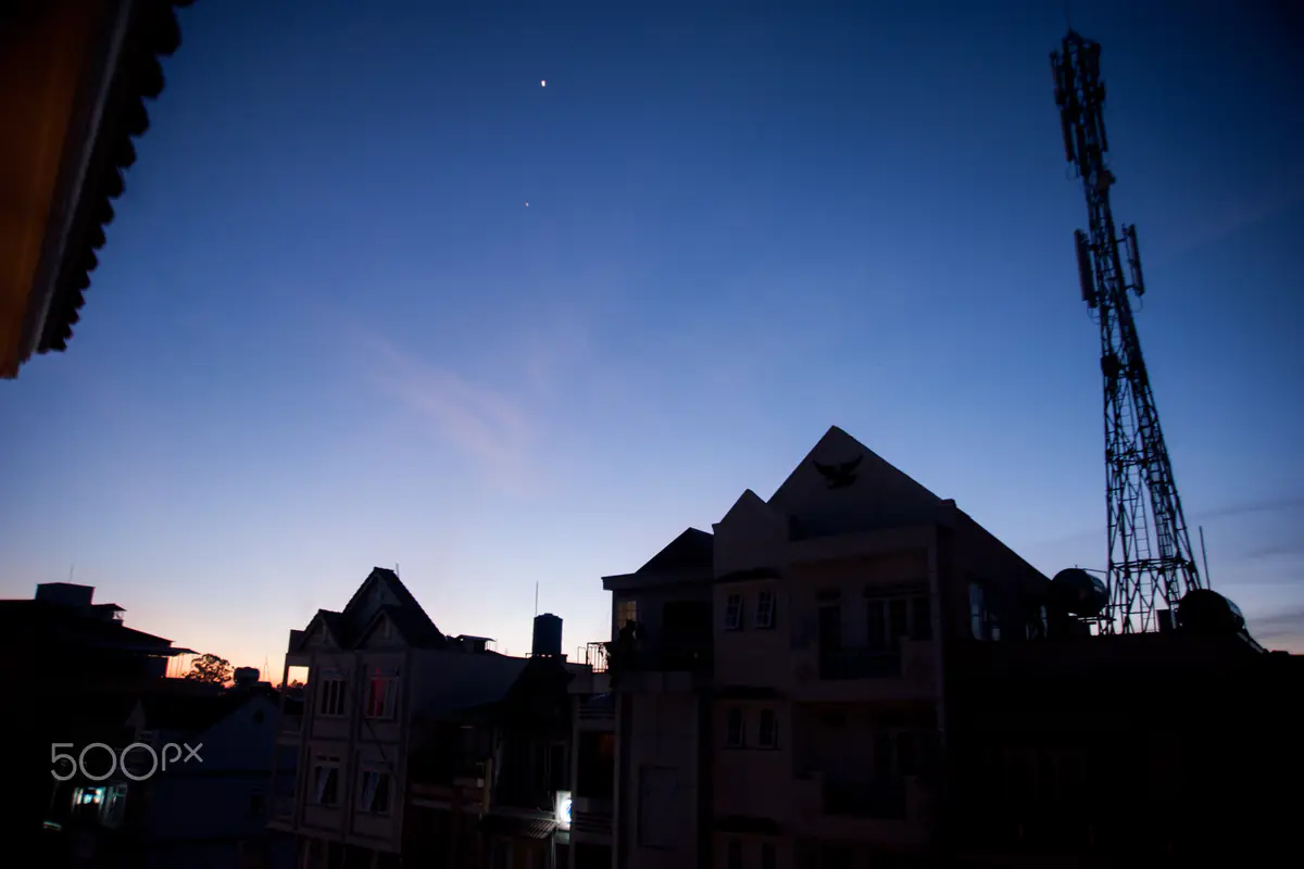 The sky over Da Lat turning from deep blue to orange at dawn, with rooftops and a telecom tower in silhouette