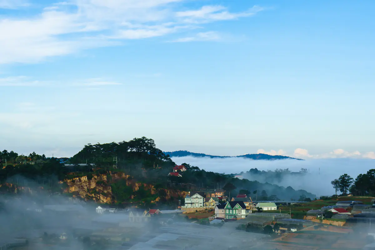 Mist wrapping a pine-covered hill and rooftops on the edge of Da Lat in the early morning, under a clear blue sky