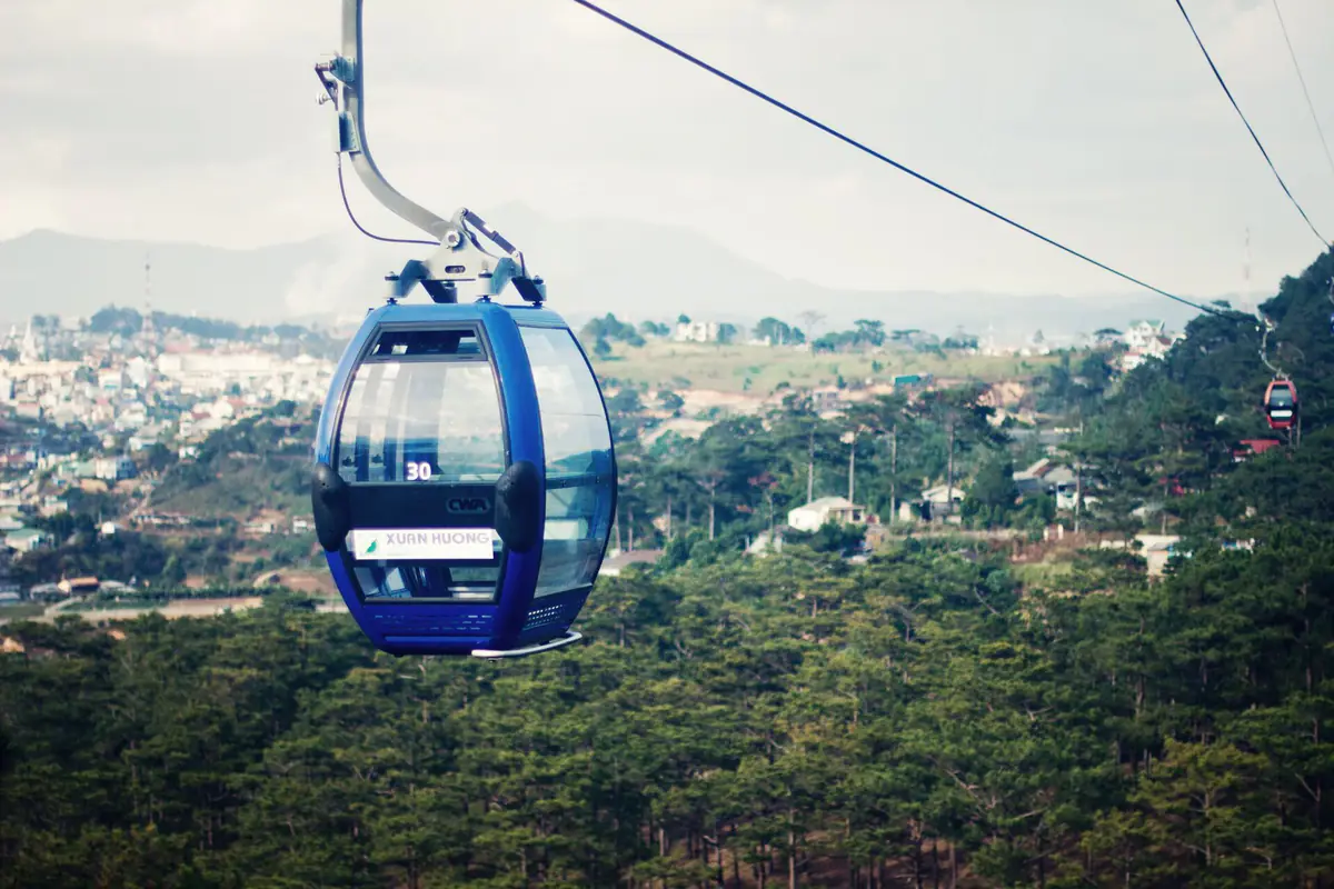 Cabin cáp treo Đà Lạt màu xanh lướt trên rừng thông, phía xa là nhà cửa thành phố và dãy núi