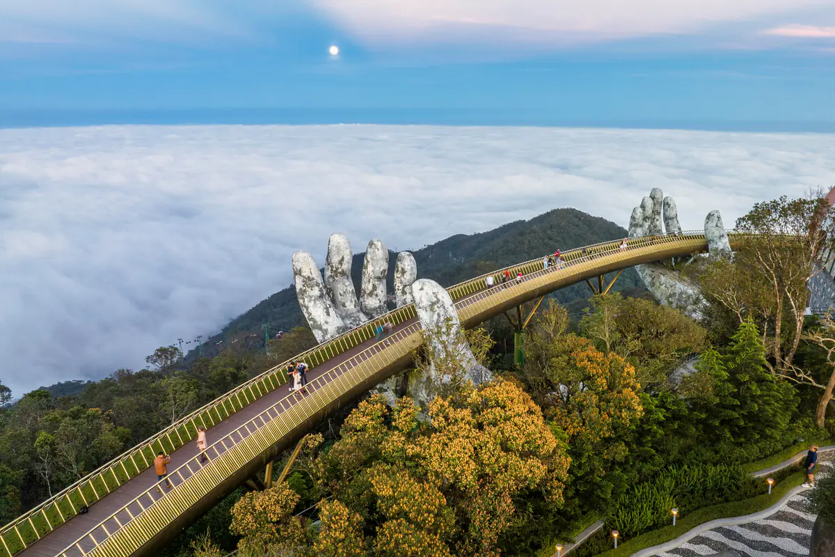 Ảnh chụp từ trên cao: Cầu Vàng và hai bàn tay đá nhô lên trên rừng, phía sau là biển mây trắng phủ kín chân núi Bà Nà dưới bầu trời xanh nhạt có vầng trăng, tháng 2/2023