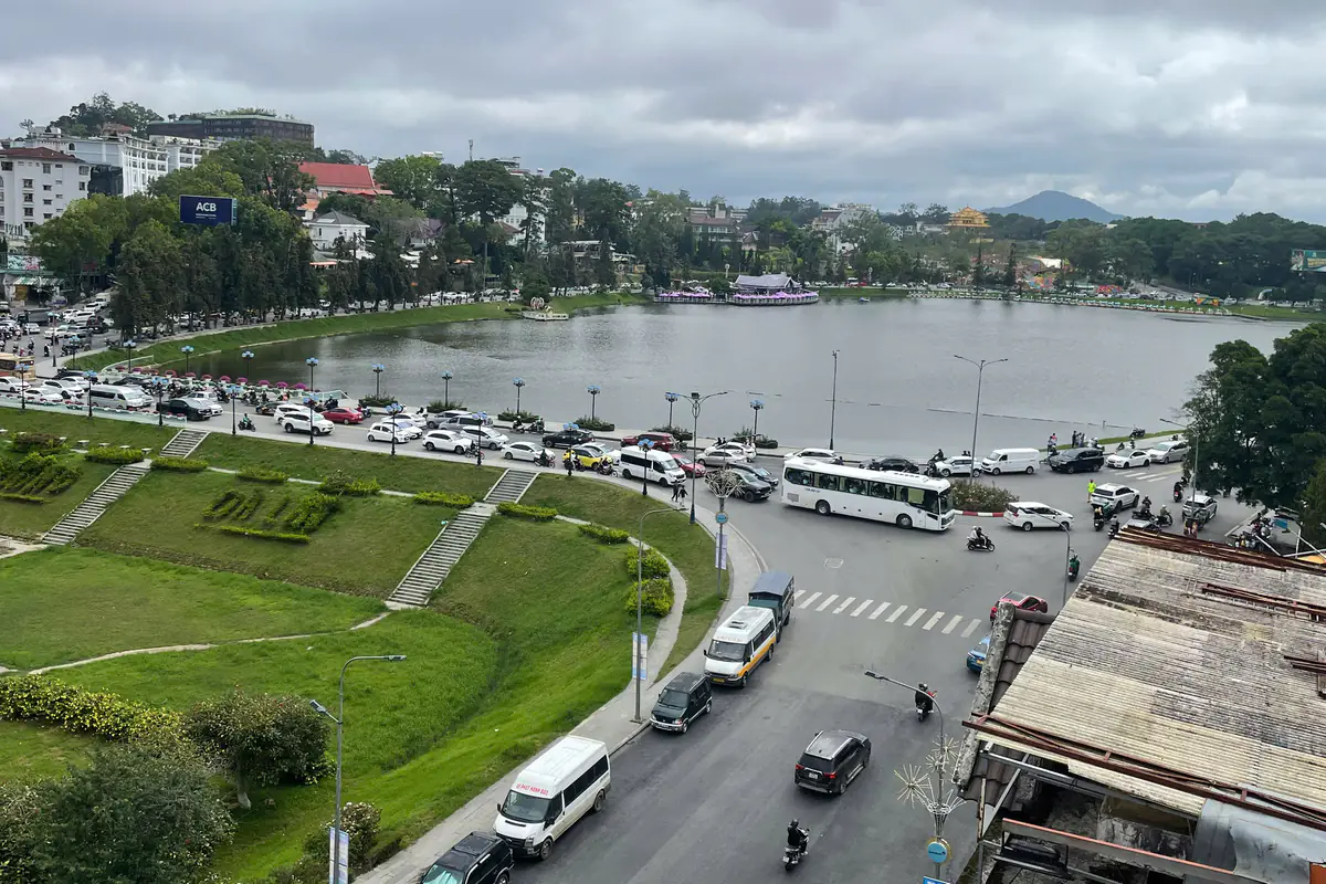 Hồ Xuân Hương và khu trung tâm Đà Lạt nhìn từ trên cao, bãi cỏ dốc, vòng xoay đông xe, đồi nhà phía xa và một ngọn núi ở chân trời