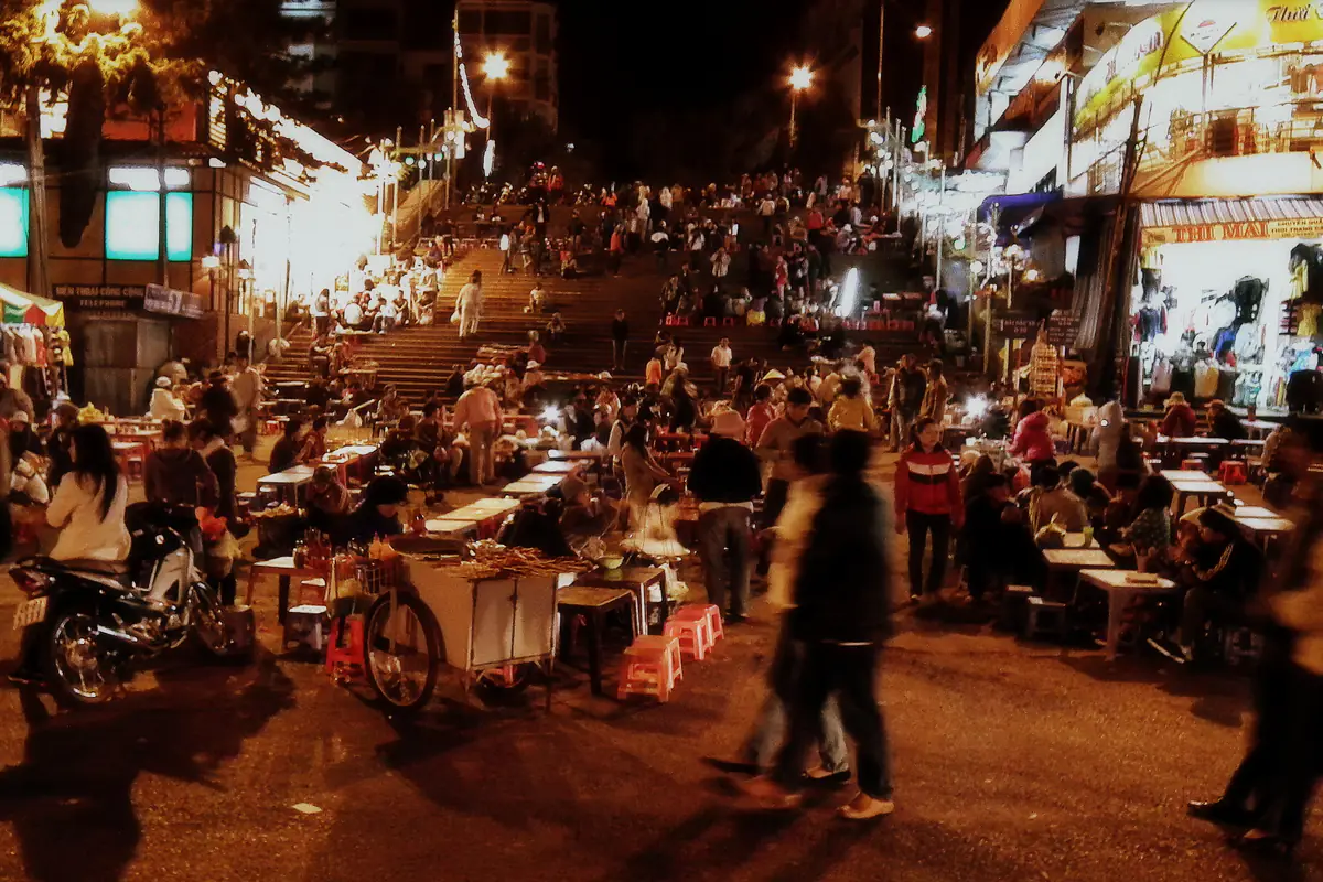 Địa điểm ăn uống Đà Lạt về đêm: hàng quán ăn vặt với bàn thấp, ghế nhựa và xe đẩy đồ nướng dưới chân bậc thang khu chợ Đà Lạt