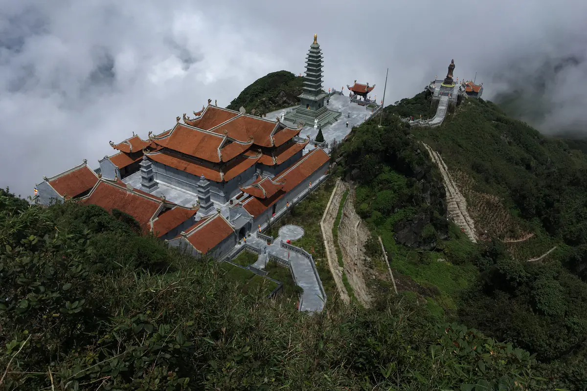 Quần thể chùa mái đỏ, bảo tháp đá và tượng Phật trên sườn núi gần đỉnh Fansipan nhìn từ trên cao, mây cuộn phía sau, tháng 8/2018