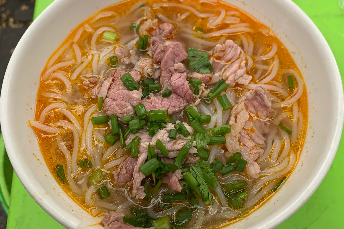 Tô bún bò Huế nước dùng màu cam đỏ, sợi bún to, thịt bò thái mỏng và nhiều hành lá, đặt trên bàn nhựa xanh ở chợ Đông Ba, tháng 7/2024