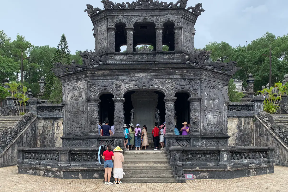 Nhà bia bằng bê tông xám sẫm, chạm trổ cầu kỳ ở lăng Khải Định, du khách đứng dưới vòm cửa và trên bậc thềm, tháng 7/2024