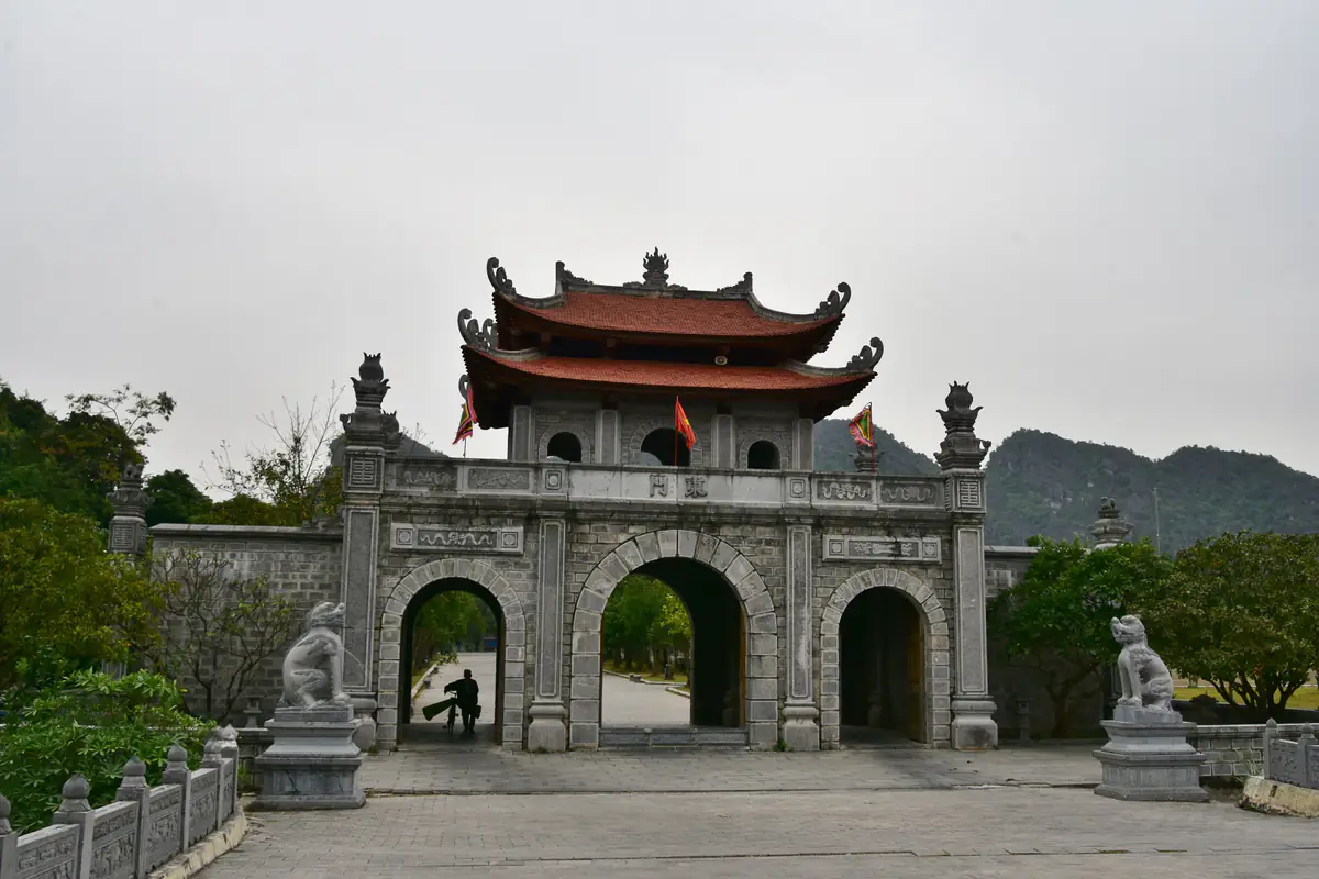 Cổng đá ba cửa vòm có mái ngói đỏ ở khu di tích Cố đô Hoa Lư, hai bên là tượng nghê đá, núi đá vôi phía sau dưới trời xám, tháng 11/2017