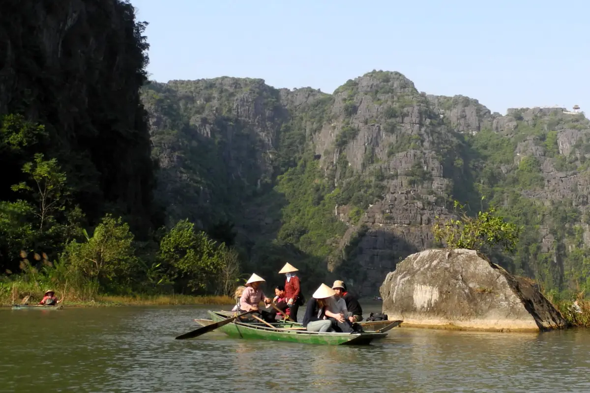 Đò chở khách đội nón lá trên sông Ngô Đồng ở Tam Cốc, cạnh một tảng đá lớn, phía sau là dãy núi đá vôi dưới trời nắng, tháng 12/2011
