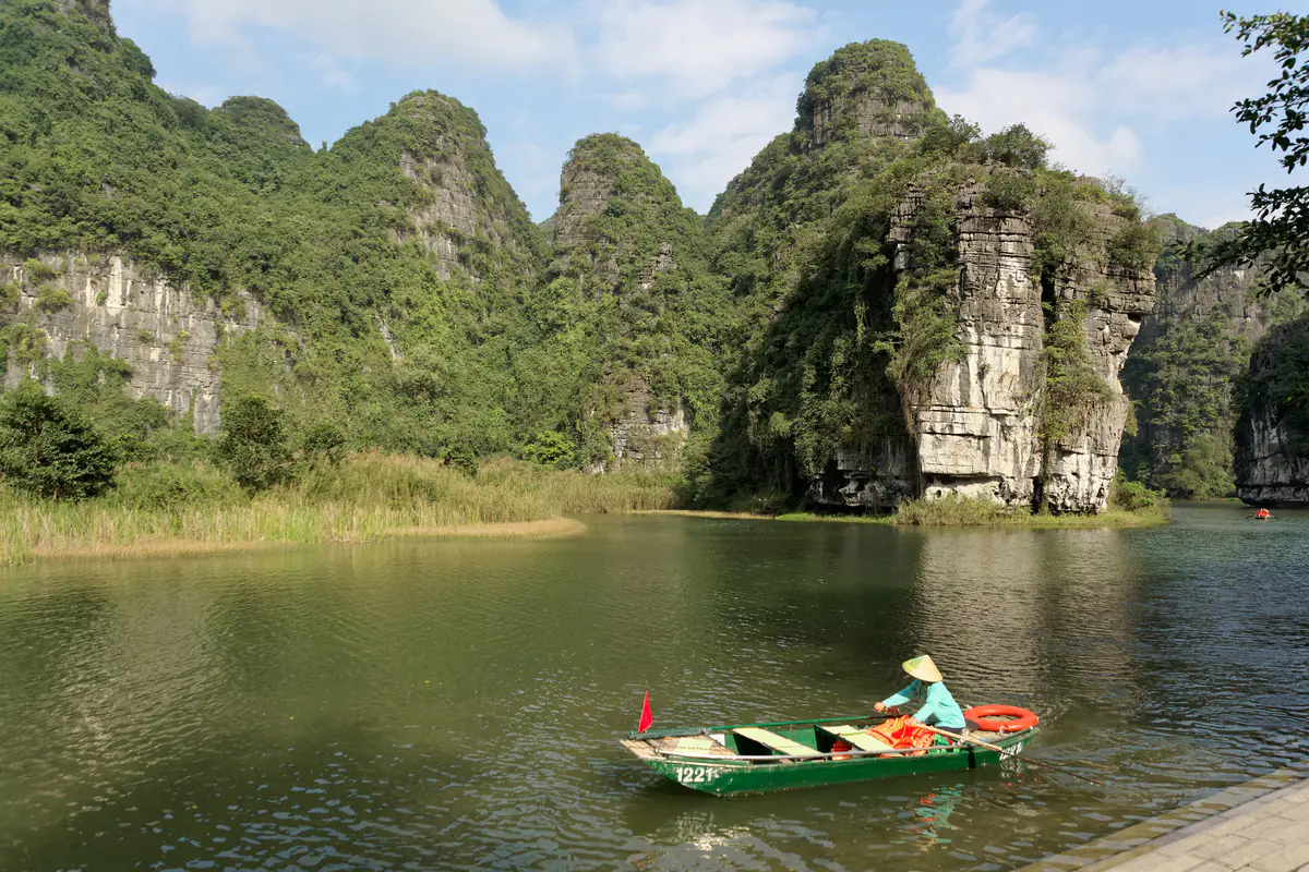 Một chiếc đò xanh do người chèo đội nón lá đi trên mặt nước Tràng An, phía sau là các khối núi đá vôi phủ cây xanh và vách đá trắng, trời nắng có mây, tháng 2/2024