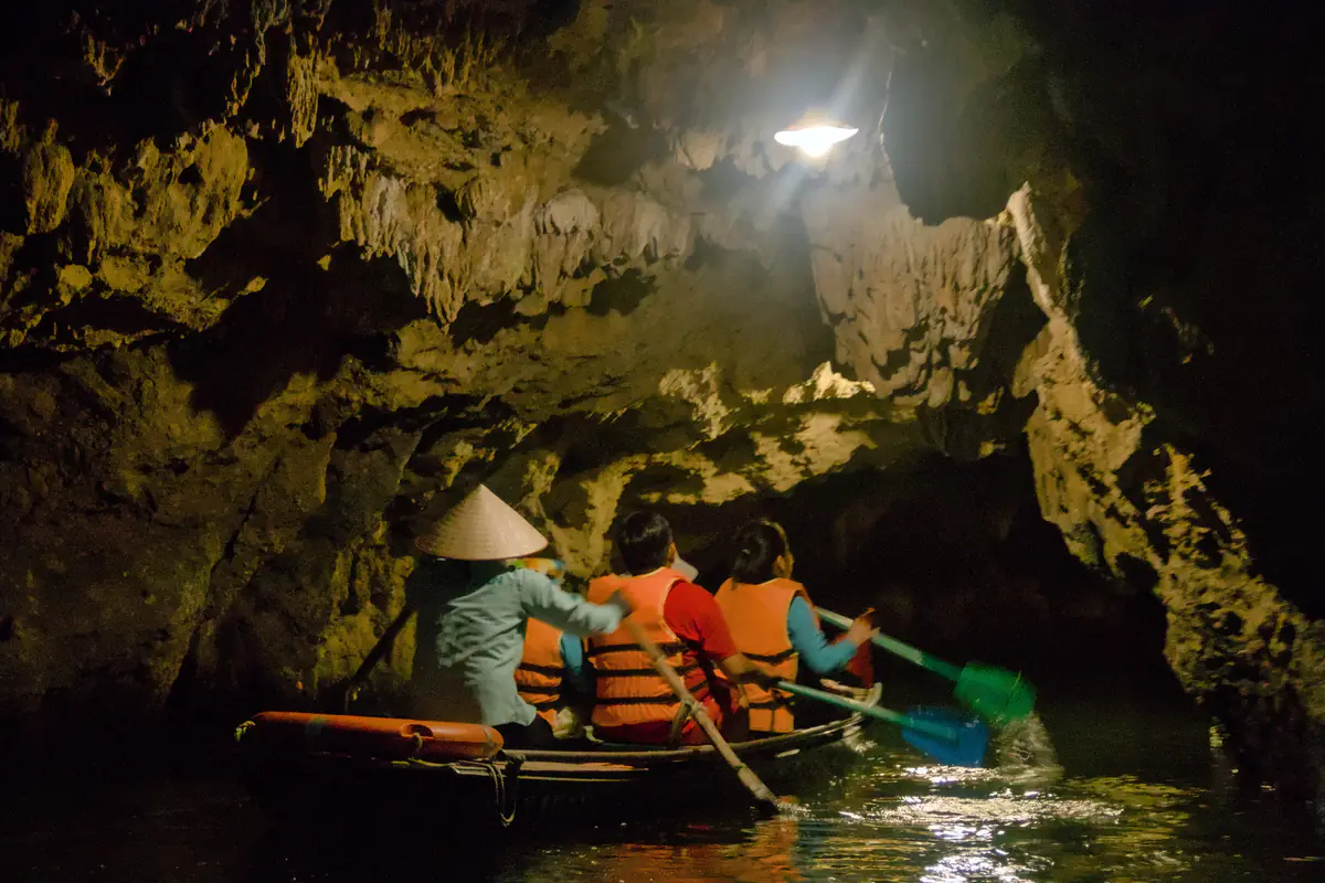 Đò chở khách mặc áo phao cam đi qua lòng một hang nước thấp ở Tràng An, trần hang lởm chởm thạch nhũ được chiếu bằng một bóng đèn, tháng 4/2023