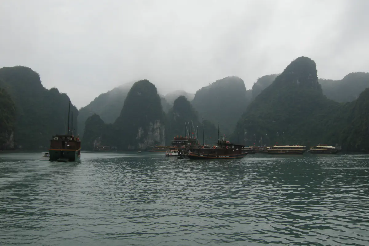 Những đảo đá vôi trên vịnh Hạ Long chìm trong sương mù buổi sáng, đỉnh núi lẫn vào nền trời xám, mặt nước gợn sóng màu xanh xám, một tàu du lịch vỏ gỗ đang chạy ra xa và vài chiếc khác neo dưới chân vách đá, sáng tháng 3/2008