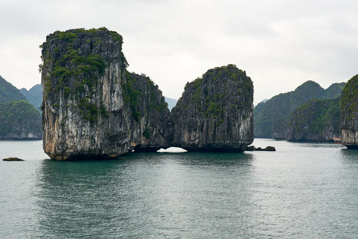 Hai khối đá vôi dựng đứng nối liền ở chân, chừa ra một vòm nhỏ sát mặt nước trên vịnh Lan Hạ gần đảo Cát Bà, cây bụi phủ xanh trên đỉnh, mặt nước xanh lam và các đảo đá khác phía sau dưới bầu trời u ám, sáng tháng 11/2023