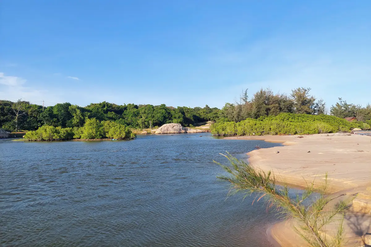 Đầm nước và doi cát ven biển Hồ Cốc, bờ có đá tảng và cây bụi xanh, trời trong vào tháng 11/2024