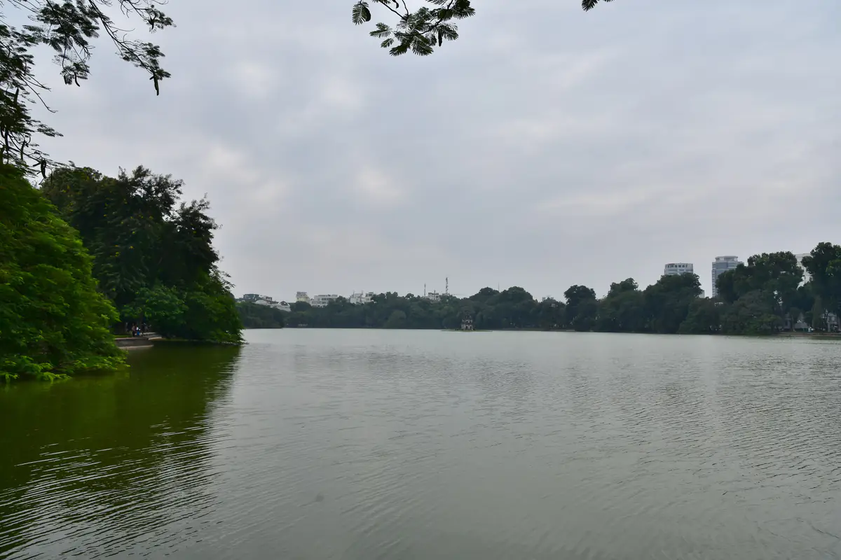 Mặt hồ Hoàn Kiếm phẳng lặng dưới bầu trời xám đục, hàng cây xanh bao quanh bờ hồ, Tháp Rùa nhỏ ở giữa xa và vài tòa nhà cao phía sau, một buổi trưa nhiều mây tháng 11/2017