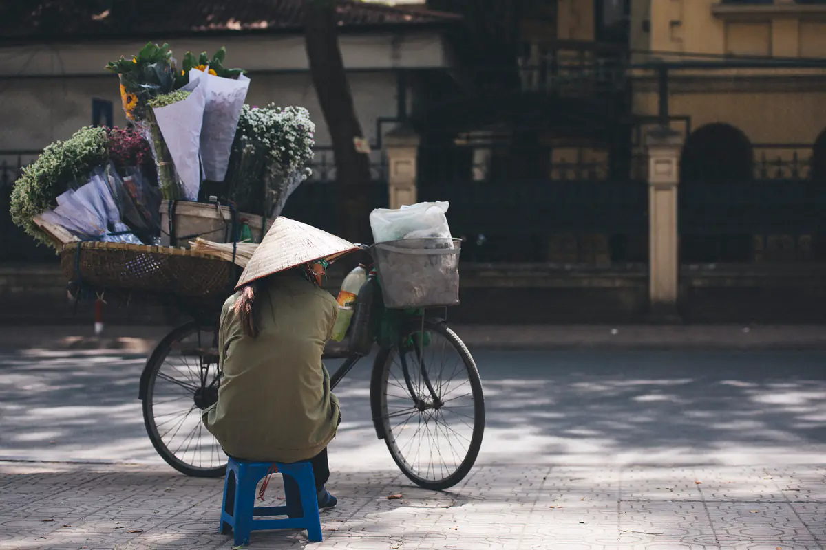 Người bán hoa đội nón lá ngồi trên ghế nhựa xanh cạnh chiếc xe đạp chở đầy bó hoa gói giấy, trên vỉa hè lát gạch rợp bóng cây ở Hà Nội, tháng 2/2017