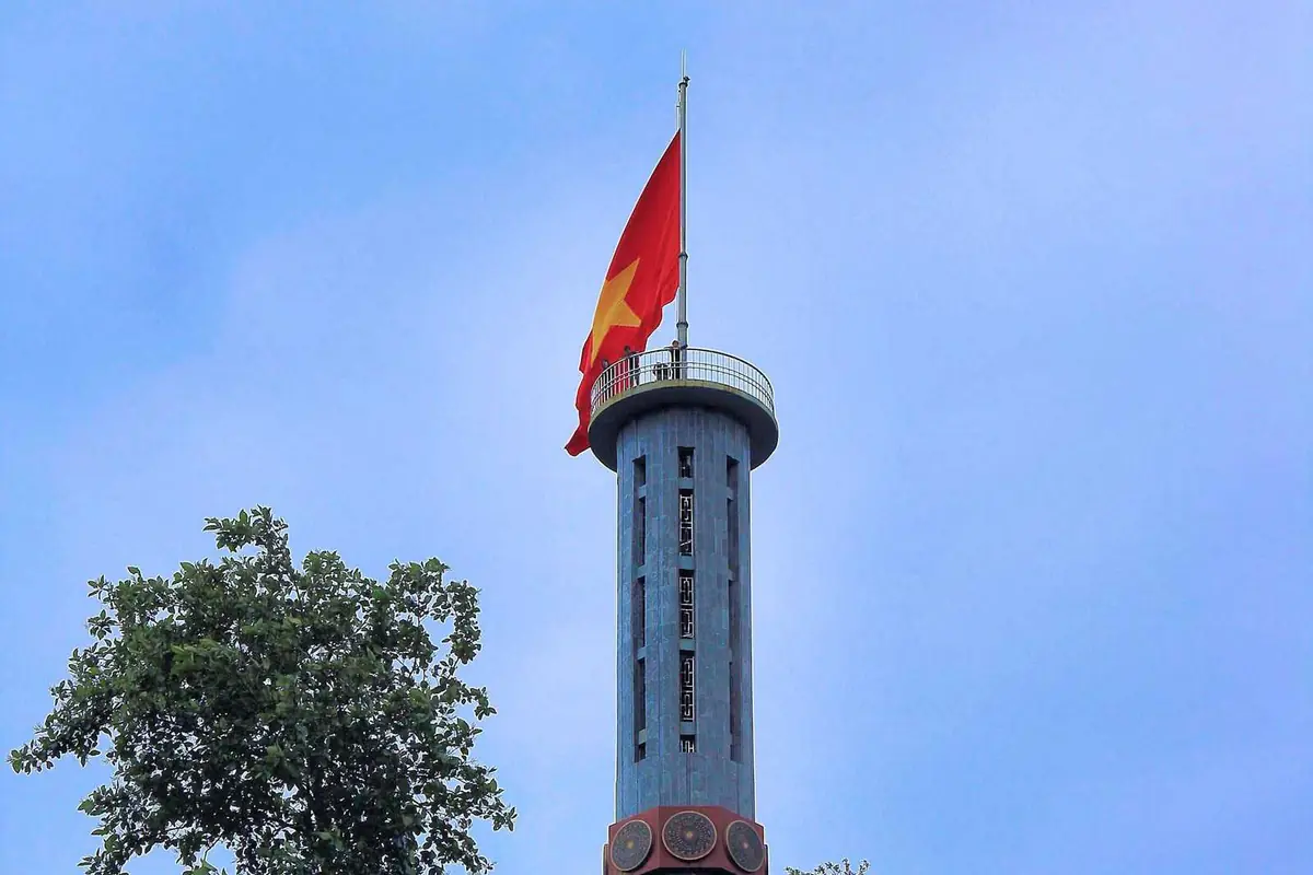 Lá cờ đỏ sao vàng bay trên đỉnh cột cờ Lũng Cú hình trụ, chân tháp có trống đồng, trên nền trời xanh, tháng 9/2013