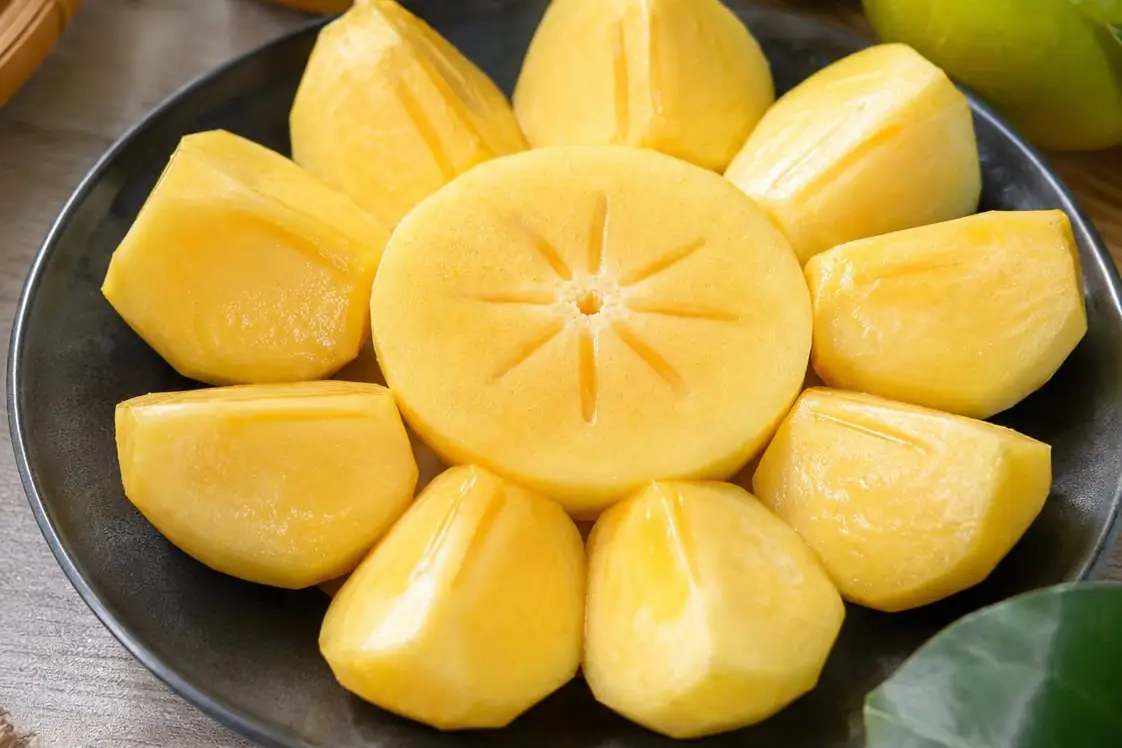Một quả hồng giòn cắt thành tám miếng xếp hình bông hoa trên đĩa đen, ruột vàng chắc, phía sau là vài quả hồng nguyên