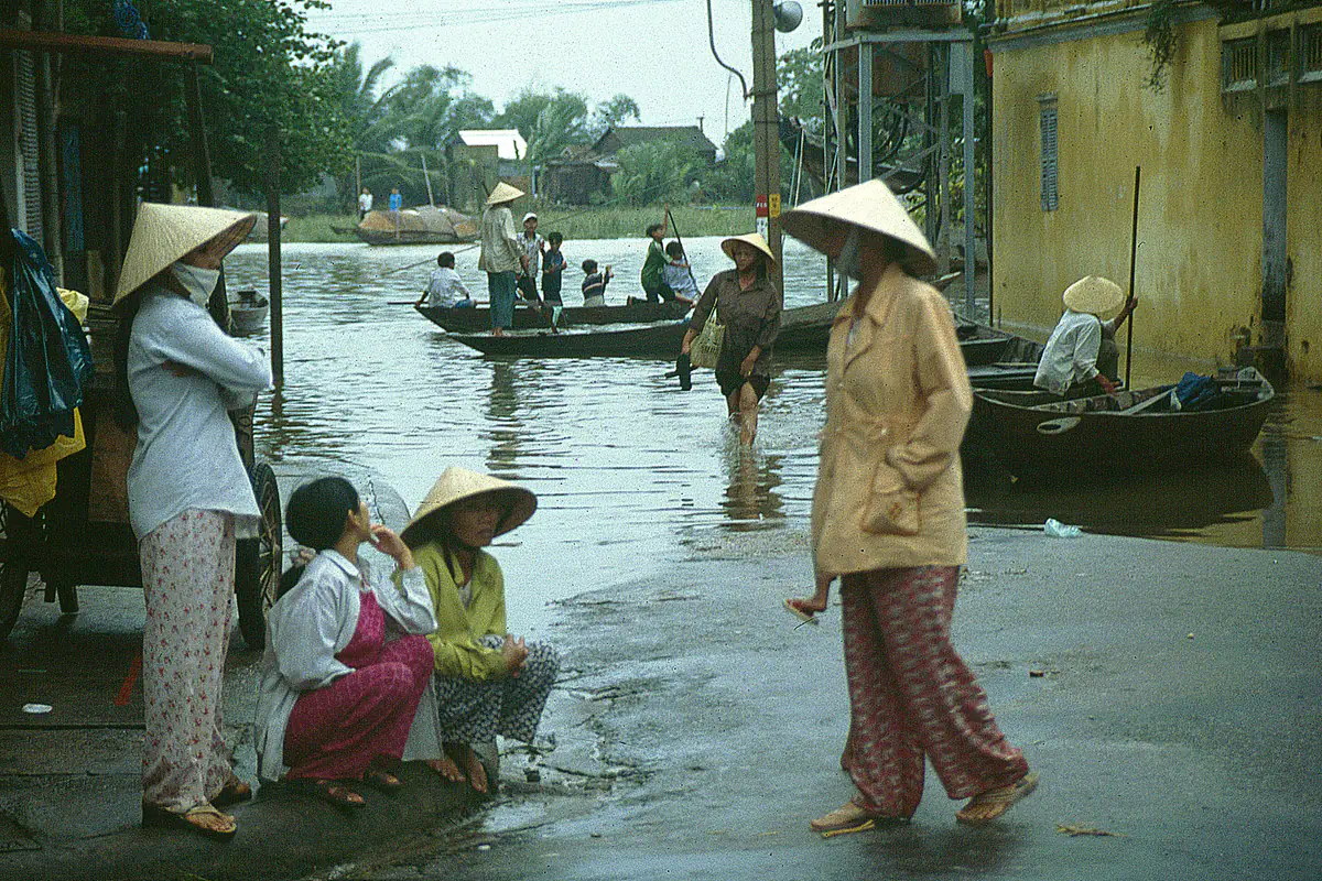 Mấy người phụ nữ đội nón lá đứng bên mép nước trên một con phố ngập ở Hội An, ghe xuồng chèo ngay giữa lòng đường trước dãy nhà tường vàng, tháng 11/2000