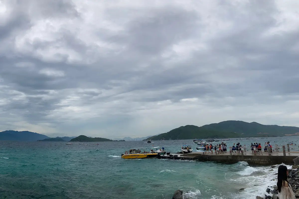Bến tàu trên đảo Hòn Mun dưới bầu trời xám nhiều mây, ca nô vàng và thuyền đậu sát cầu tàu đông khách, sóng vỗ vào kè đá, phía xa là Hòn Một, Hòn Tằm và Hòn Tre, tháng 7/2026