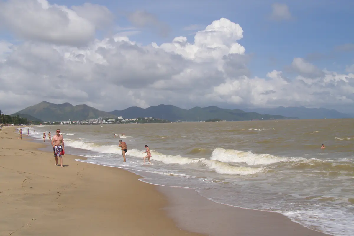Bãi biển Nha Trang một buổi sáng tháng 11/2013, nước biển đục màu nâu, sóng bạc đầu nối nhau vào bờ, vài người lội nước ở mép sóng, phía sau là dãy núi và nhà cao tầng dưới trời nhiều mây