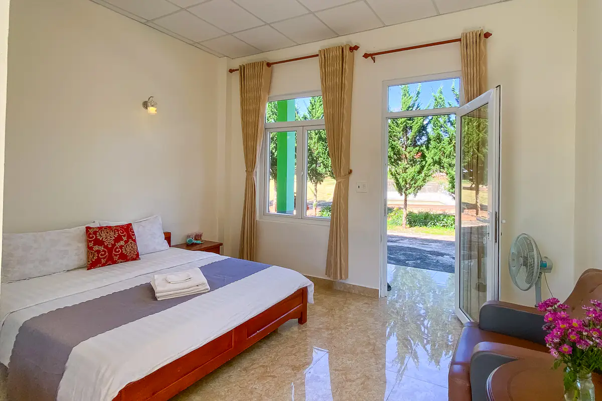 Phòng studio có giường đôi trải ga trắng và gối đỏ, cửa sổ, cửa kính mở ra lối đi có hàng cây, ghế bành và quạt đứng ở Robins Farmstay, Tà Nung, Đà Lạt