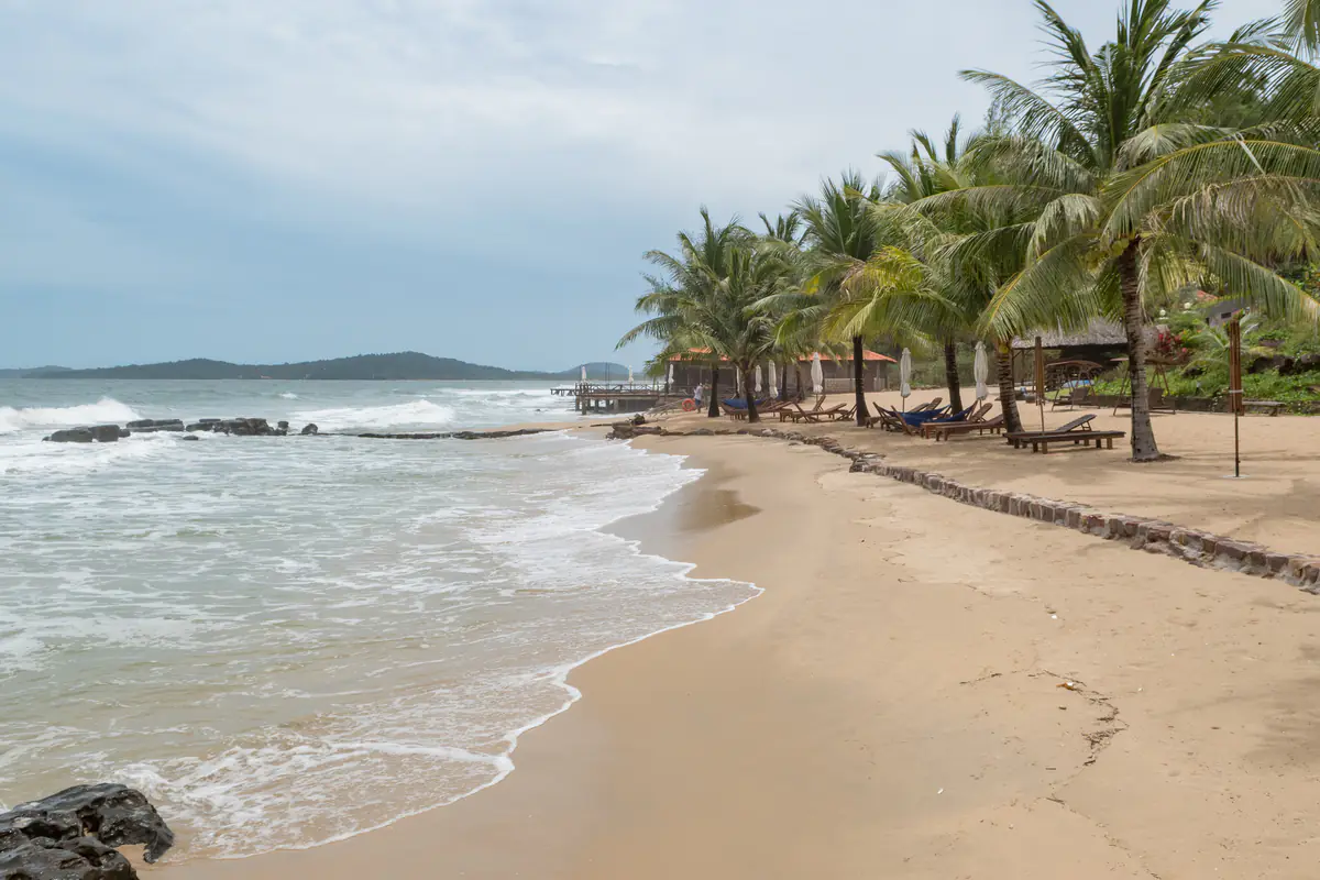 Bãi biển cát vàng cong theo mép nước ở khu vực Ông Lang, bờ tây Phú Quốc phía bắc Dương Đông, sóng bạc đầu vỗ vào ghềnh đá, hàng dừa nghiêng cùng ghế tắm nắng bằng gỗ và một cầu gỗ nhỏ phía xa, buổi sáng nhiều mây tháng 8/2017
