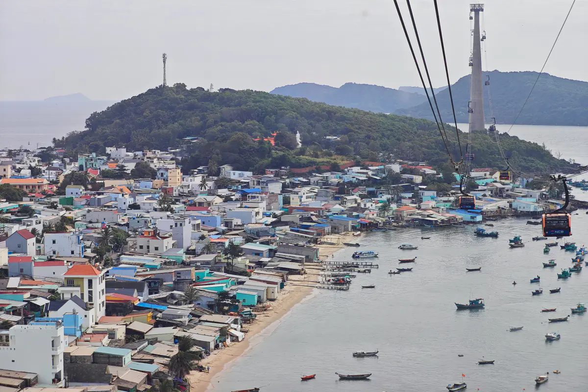 Khu dân cư ven biển An Thới nhìn từ cabin cáp treo Hòn Thơm, với dãy nhà san sát bên bãi cát, nhiều thuyền đánh cá neo trong vịnh, trụ cáp bê tông cùng các cabin trên đường cáp và các đảo thuộc quần đảo An Thới phía xa, trưa tháng 12/2025
