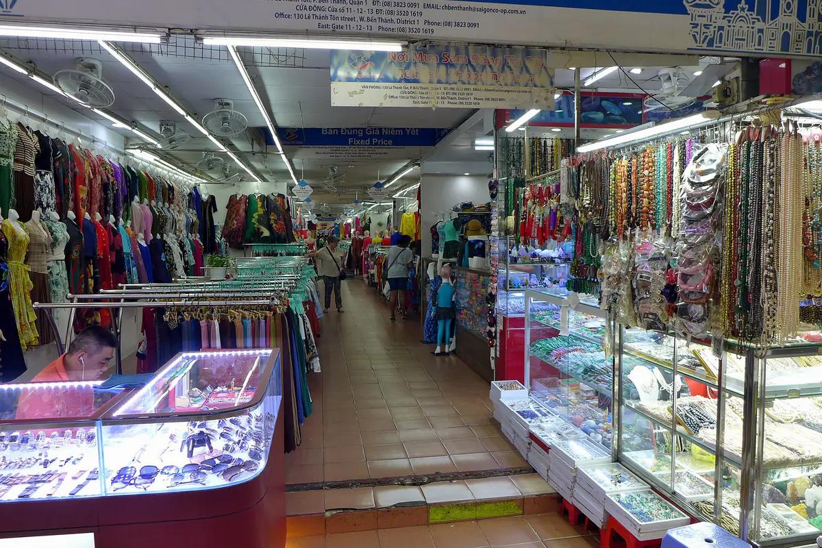 Rows of clothing, fabric and jewellery stalls in Ben Thanh Market, November 2023, under a blue overhead sign reading “Bán đúng giá niêm yết - Fixed Price”