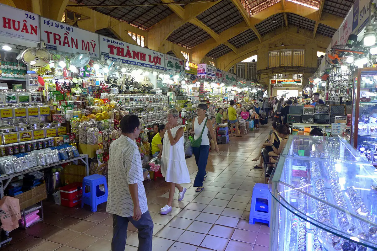 An aisle inside Ben Thanh Market, Ho Chi Minh City, November 2023: stalls on the left are stacked with packaged coffee, cashews, candied and dried fruit, tourists walk down the aisle and a glass jewellery counter stands on the right
