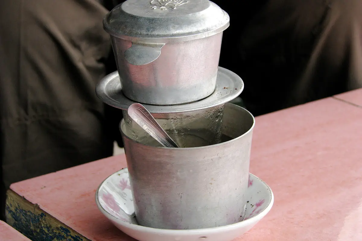 An aluminium phin filter sitting on an aluminium cup and a china saucer on a pink stone table, with coffee dripping through, December 2008