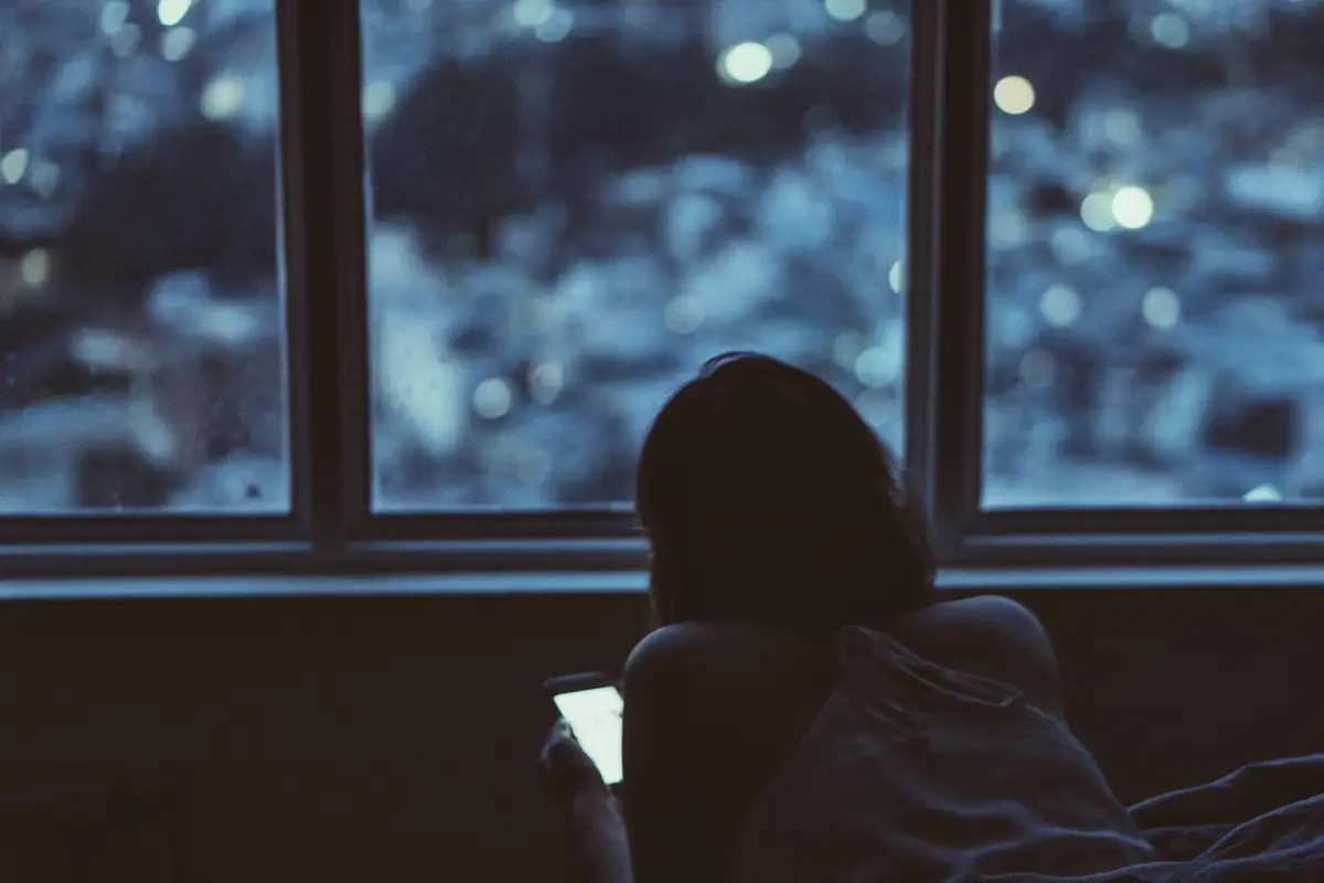 Một người nằm nhìn màn hình điện thoại sáng trong phòng tối, sau cửa kính là đèn thành phố mờ nhòe, TP.HCM, tháng 10/2016