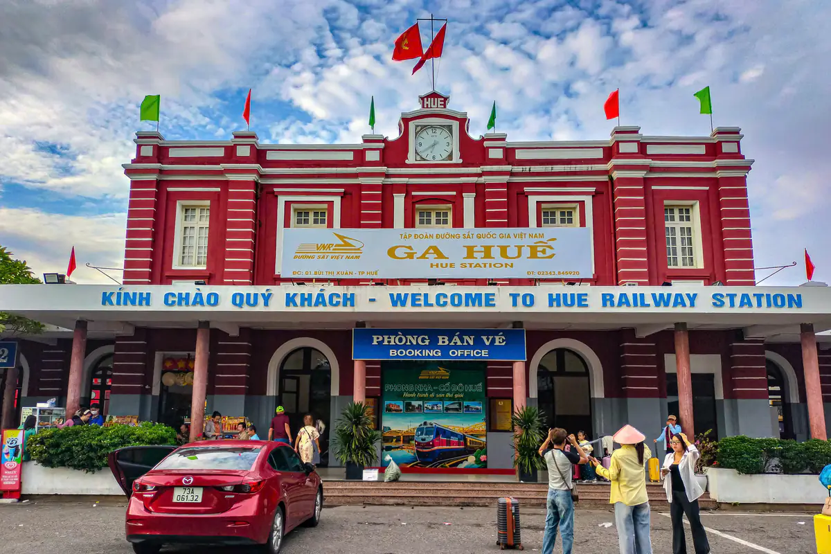 Mặt tiền ga Huế sơn đỏ với tháp đồng hồ, biển Kính chào quý khách - Welcome to Hue Railway Station và lối vào phòng bán vé, hành khách đứng chụp ảnh trước sân ga sáng 2/9/2026