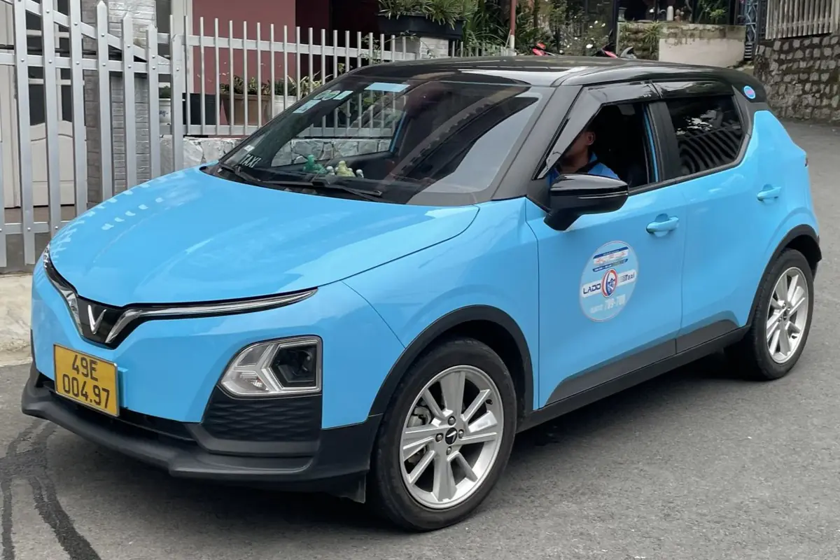 Taxi điện VinFast VF 5 màu xanh da trời của hãng Lado trên một con đường dốc ở Đà Lạt, biển số nền vàng, logo hãng dán trên cửa trước