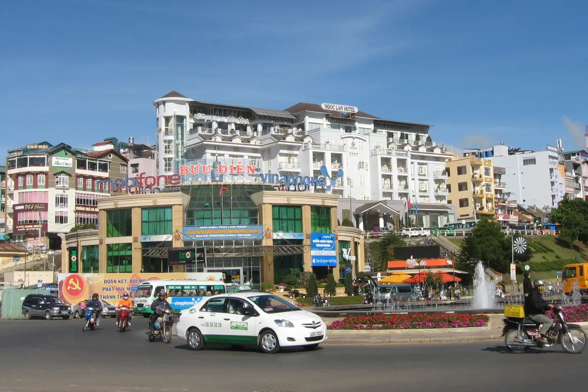 Taxi Mai Linh màu trắng xanh chạy qua vòng xoay trước Bưu điện trung tâm Đà Lạt năm 2011, phía sau là khách sạn Ngọc Lan và xe máy