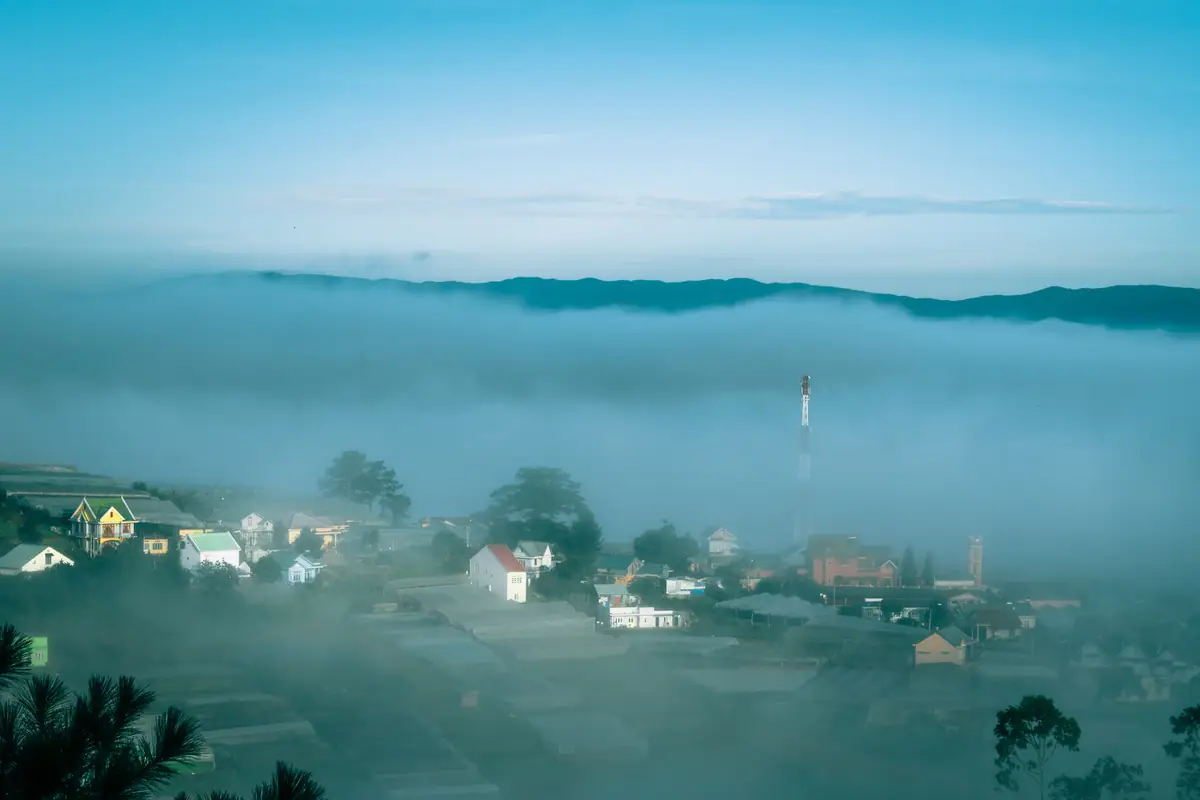 Early-morning fog filling the Trai Mat valley on the edge of Da Lat, with rooftops and a radio mast poking through and a mountain ridge behind