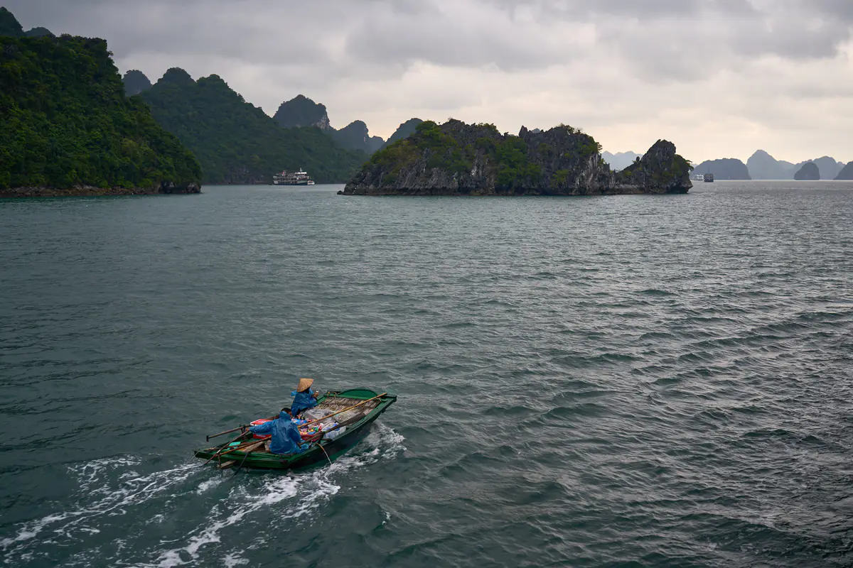 Chiếc thuyền nhỏ của người bán hàng trên vịnh Hạ Long rẽ sóng dưới bầu trời u ám, phía sau là các đảo đá vôi và một tàu du lịch, tháng 11/2023