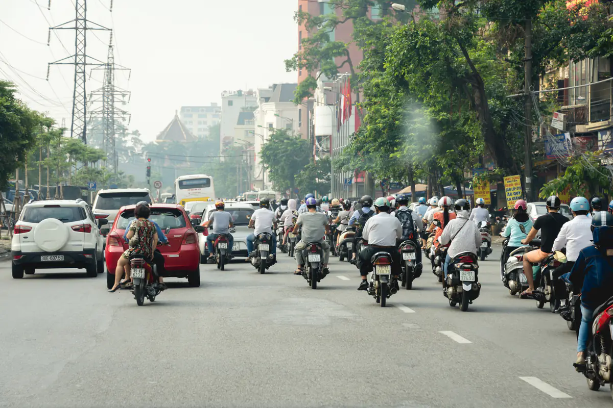 Dòng xe máy và vài chiếc ô tô cùng chạy trên một con đường thành phố ở Việt Nam, người lái đội mũ bảo hiểm, hai bên là hàng cây và nhà phố, chụp từ trong xe