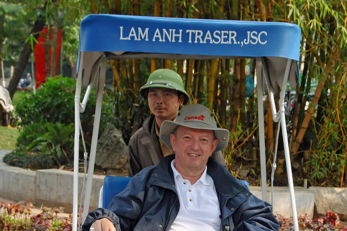 Một du khách nước ngoài đội mũ vải ngồi trên xích lô mui xanh ở Hà Nội, người đạp xích lô đội mũ cối xanh ngồi phía sau, nền là bụi tre, năm 2009