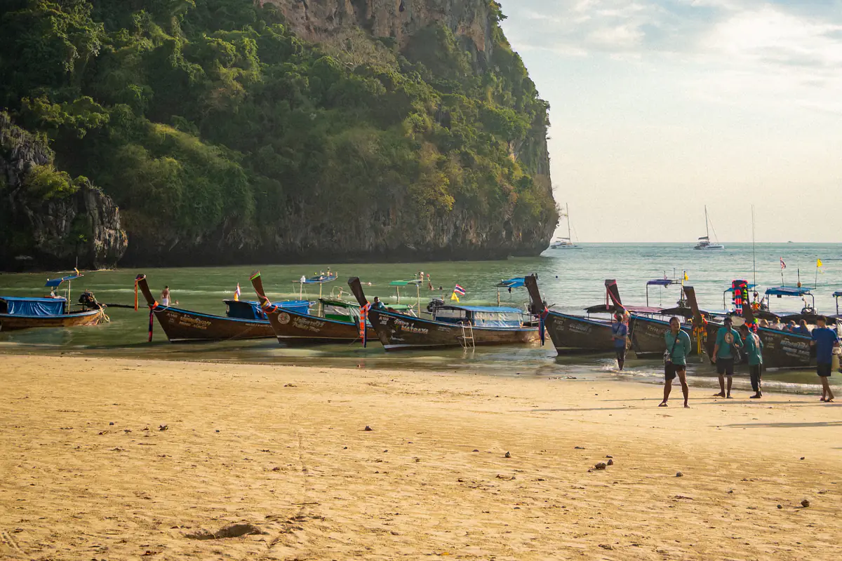 Thuyền đuôi dài neo sát mép nước ở bãi Railay Tây, Krabi, dưới chân vách núi đá vôi phủ cây xanh, vài người đứng trên bãi cát, xa xa là thuyền buồm, tháng 3/2026