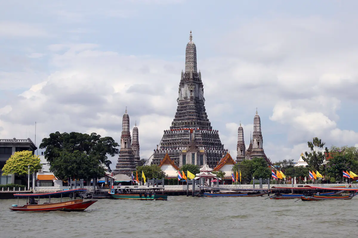 Chùa Wat Arun bên bờ sông Chao Phraya ở Bangkok, tháp chính cao vút giữa bốn tháp nhỏ, phía trước là thuyền đuôi dài và thuyền khách trên mặt sông, dãy cờ Thái và cờ vàng dọc bến, trời nhiều mây, tháng 3/2012
